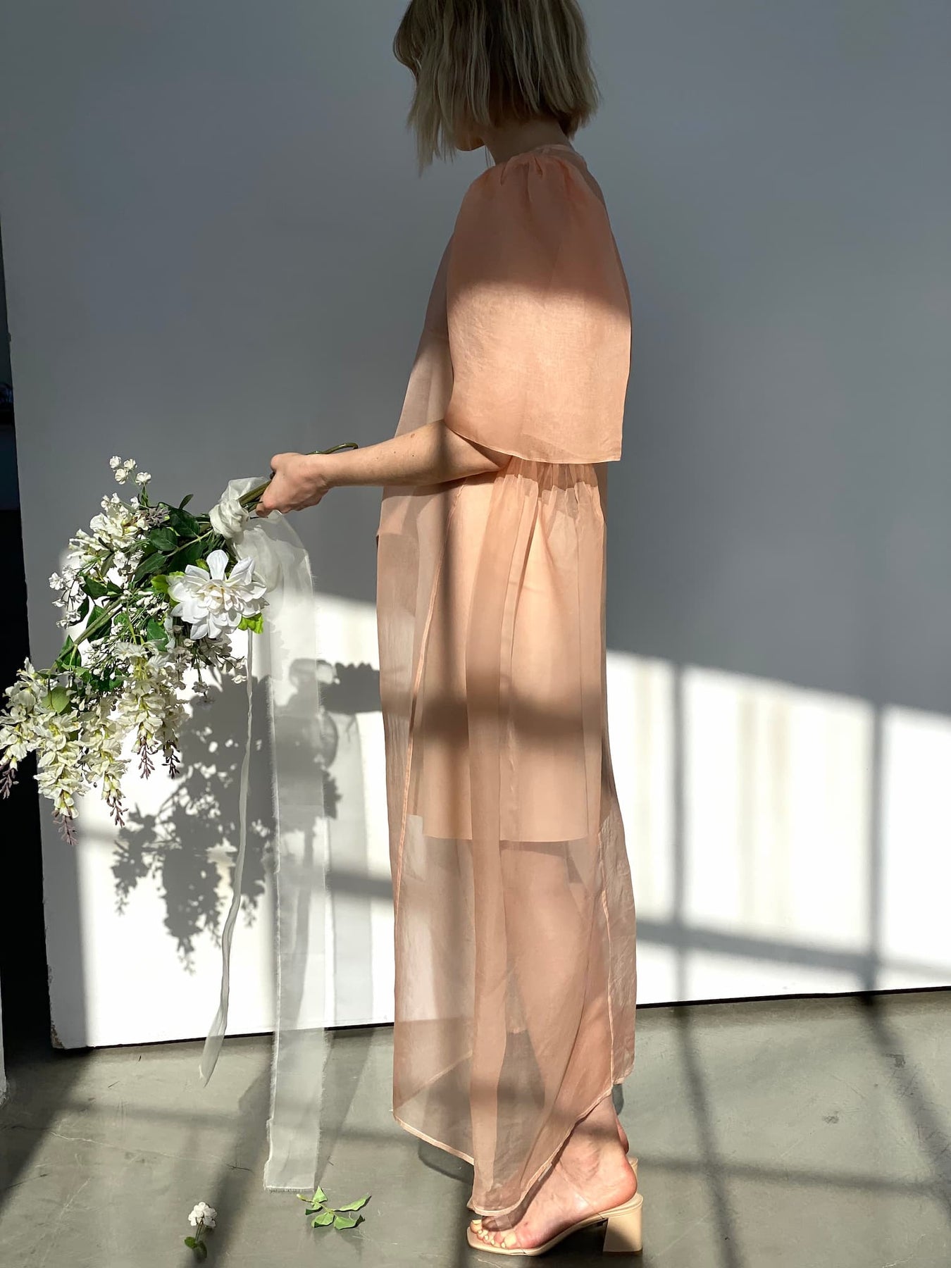 A person in a sheer peach dress stands holding a bouquet of white flowers with silk ribbons. They are in profile, with sunlight creating shadows on the wall behind them.