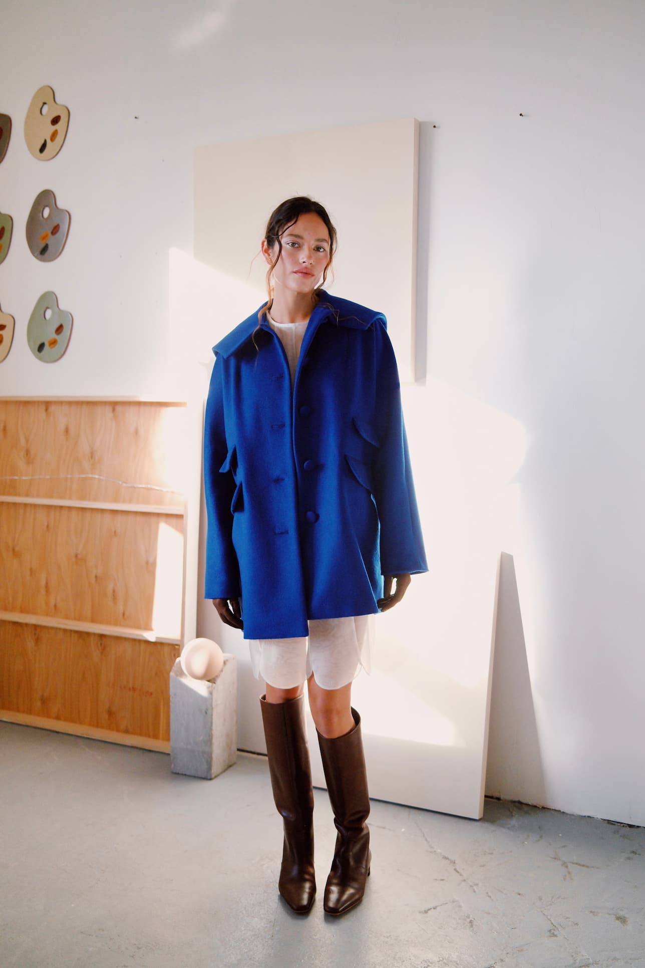 A woman stands indoors wearing a bright blue coat, white dress, and tall brown boots. She is in a minimalist room with modern art on the wall and wooden shelves in the background.