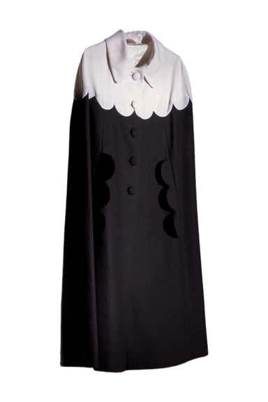 The Adèle Cape by Noémiah boasts a luxurious draped silhouette, showcasing a chic black and white wool design. It features a cape-like structure with the upper section in white, adorned with scalloped edges, and the lower section in black, echoing the refined scalloped details around the buttons. This elegant piece stands out beautifully against a neutral background.