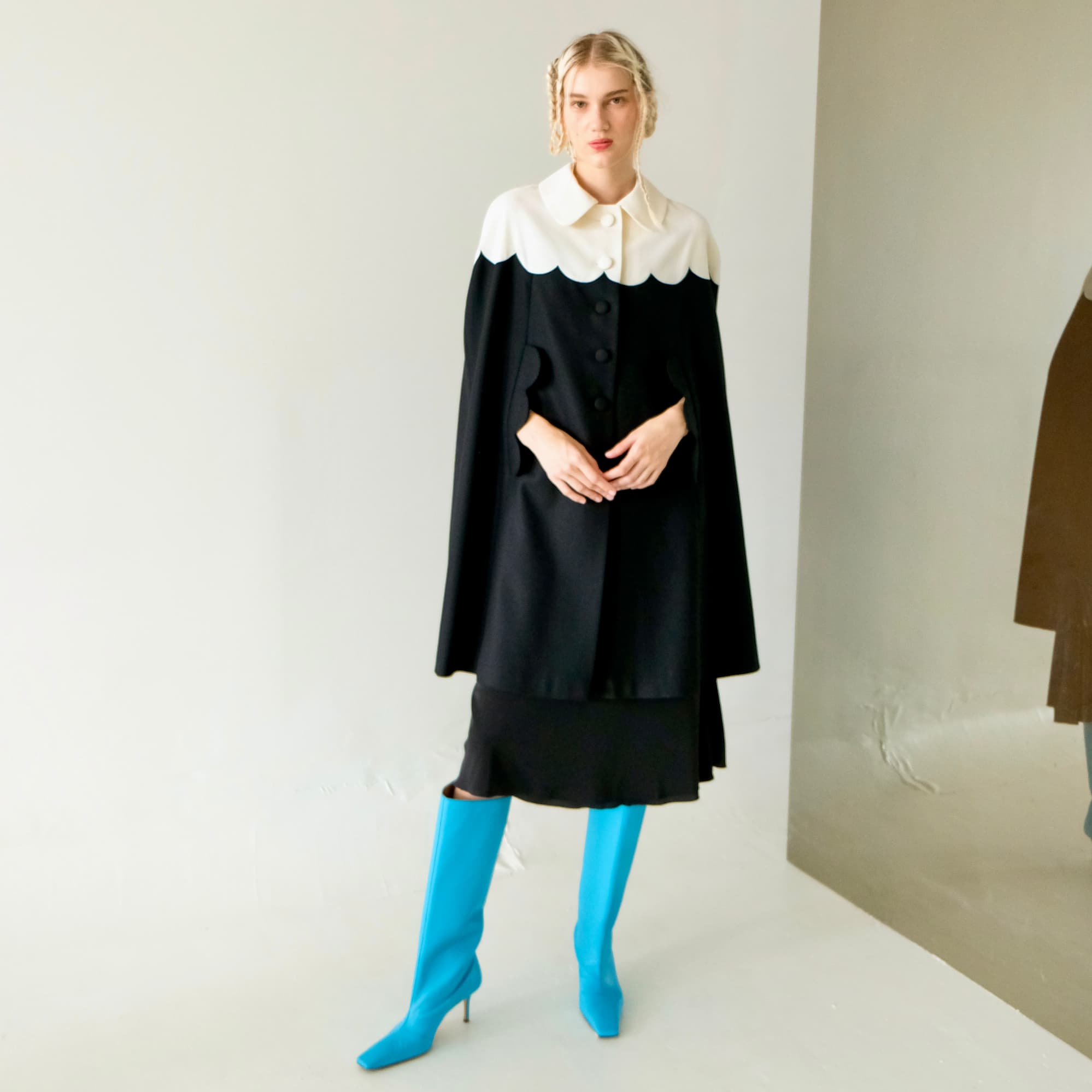 In a minimalist room, a person is elegantly dressed in the Noémiah Adèle Cape, a luxurious black garment featuring a white scalloped collar. This wool cape's draped silhouette accentuates their braided hair and harmonizes beautifully with striking blue knee-high boots. A reflective surface on the right completes the scene.