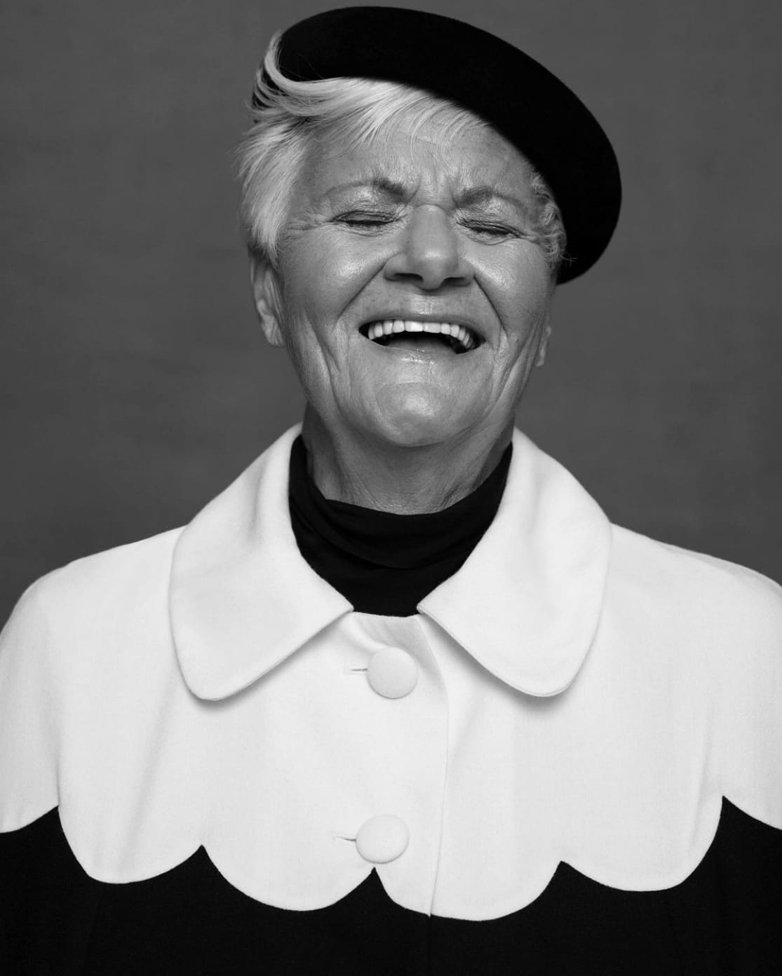 A black and white portrait of an older person with short white hair, wearing a black beret and the Adèle Cape by Noémiah, which features a scalloped collar. The draped silhouette enhances their elegance as they smile broadly with closed eyes, radiating joy.