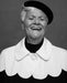 A black and white portrait of an older person with short white hair, wearing a black beret and the Adèle Cape by Noémiah, which features a scalloped collar. The draped silhouette enhances their elegance as they smile broadly with closed eyes, radiating joy.