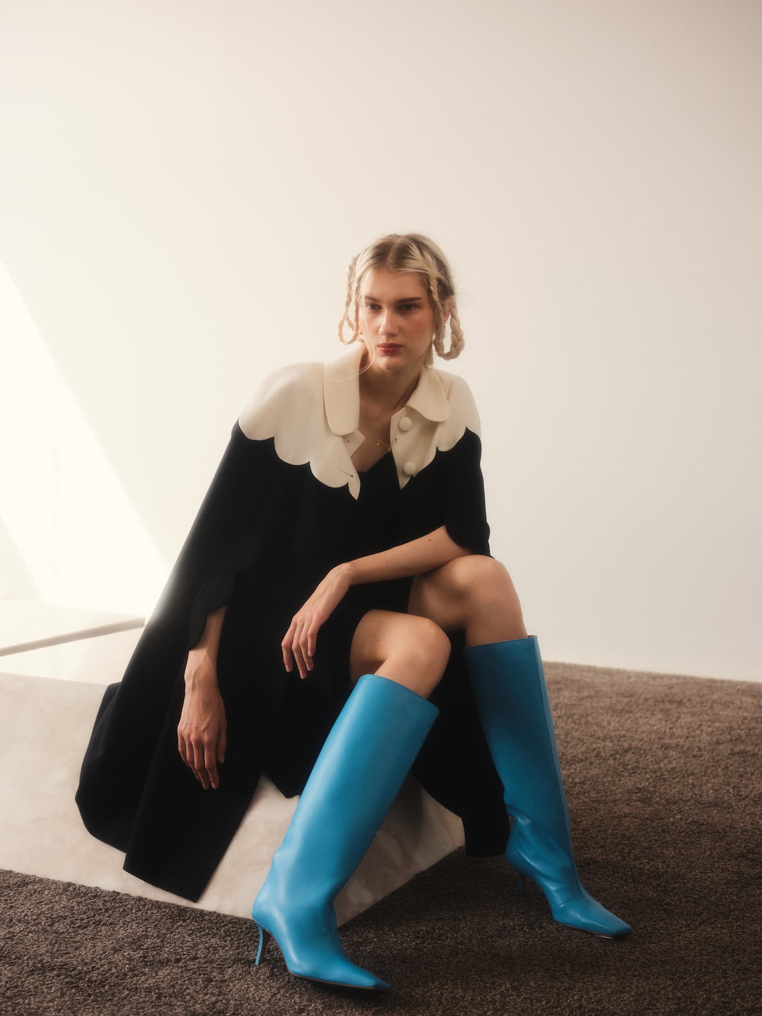 A person with braided hair is sitting on a platform in a sunlit room, wearing luxury clothing from Noémiah. Their Adèle Cape pairs elegantly with bright blue knee-high boots. The minimalist setting features brown carpet and white walls, enhancing the sophisticated ambiance.