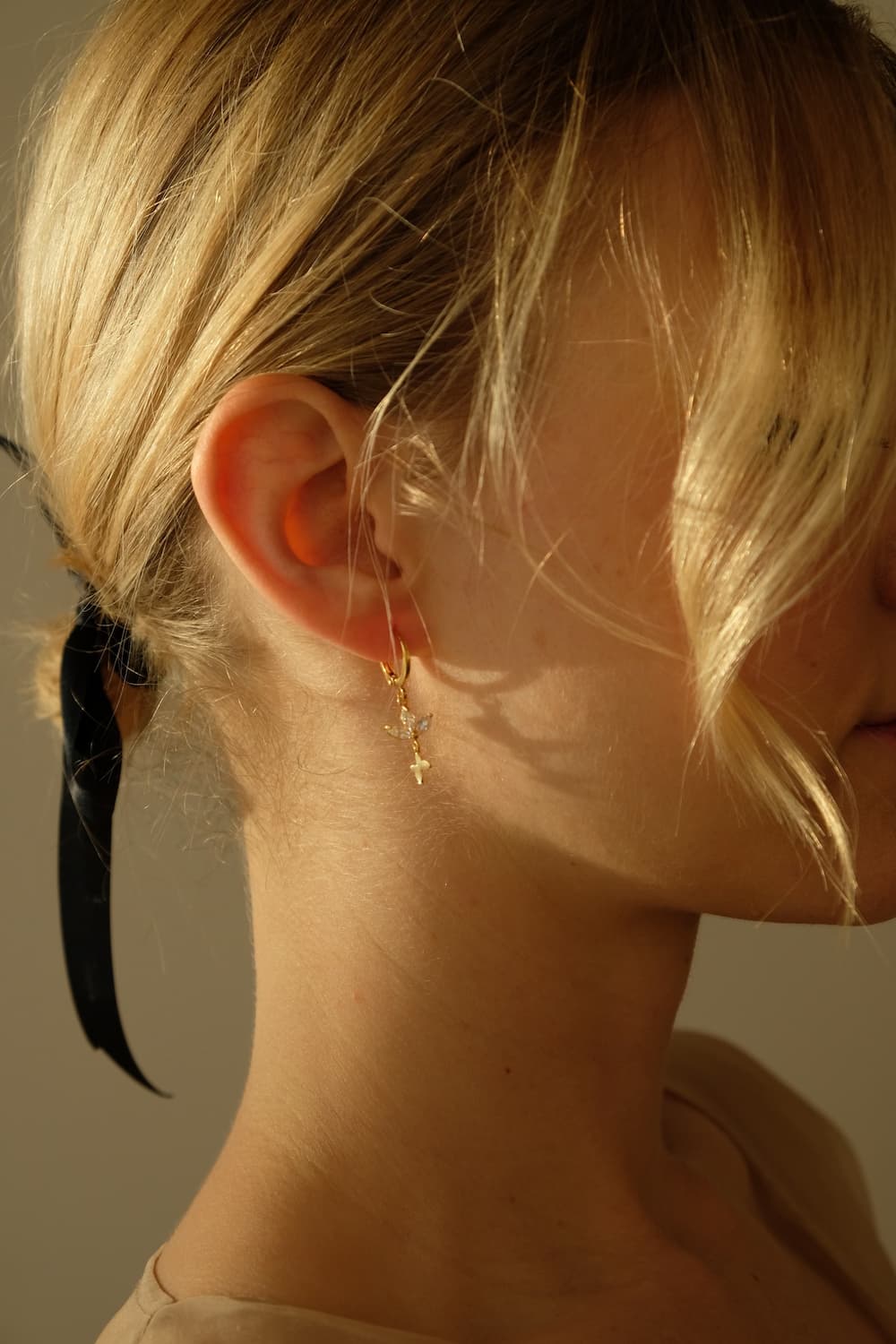 A woman with blonde hair is shown in profile, wearing a black ribbon and a pair of Alaska Earrings by Noémiah. The warm lighting highlights her elegant accessories.