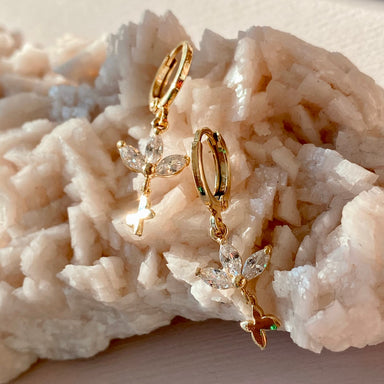 The Alaska Earrings by Noémiah are showcased on a textured, light-colored rock. These gold-plated hoop earrings feature dangling fairy charms adorned with cubic zirconia, and their delicate, translucent wings create a stunning sparkling effect as they catch the light.