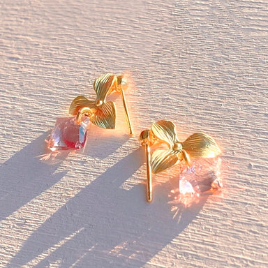 The Annette Earrings by Noémiah are a pair of tarnish-resistant gold earrings adorned with floral designs and pink Swarovski crystals. These everyday pieces beautifully catch the sunlight, casting long shadows on a textured, light-colored surface, while showcasing their delicate craftsmanship.