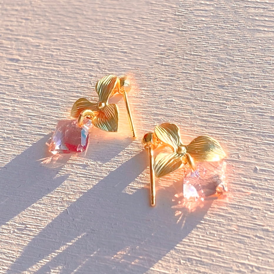 The Annette Earrings by Noémiah are a pair of tarnish-resistant gold earrings adorned with floral designs and pink Swarovski crystals. These everyday pieces beautifully catch the sunlight, casting long shadows on a textured, light-colored surface, while showcasing their delicate craftsmanship.