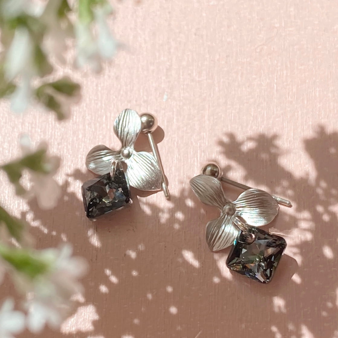 The Annette Earrings by Noémiah feature a stunning silver flower shape, adorned with dark, faceted stones and Swarovski crystals. These earrings cast delicate shadows on a light pink surface accompanied by softly blurred white flowers. Tarnish-resistant, they are ideal for adding everyday elegance.