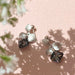 The Annette Earrings by Noémiah feature a stunning silver flower shape, adorned with dark, faceted stones and Swarovski crystals. These earrings cast delicate shadows on a light pink surface accompanied by softly blurred white flowers. Tarnish-resistant, they are ideal for adding everyday elegance.