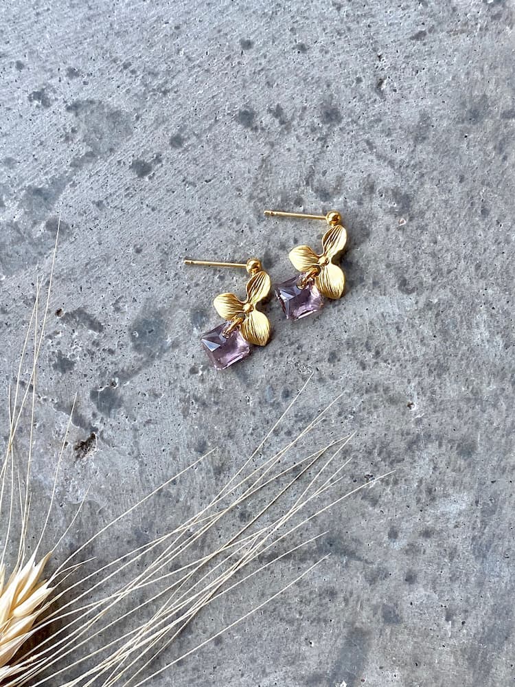 The Annette Earrings by Noémiah, featuring purple gemstones and embellished with Swarovski crystals, rest gracefully on a textured gray surface. Beside them, delicate dried plants provide a natural decorative touch. These tarnish-resistant earrings are ideal for everyday wear, blending elegance with durability.