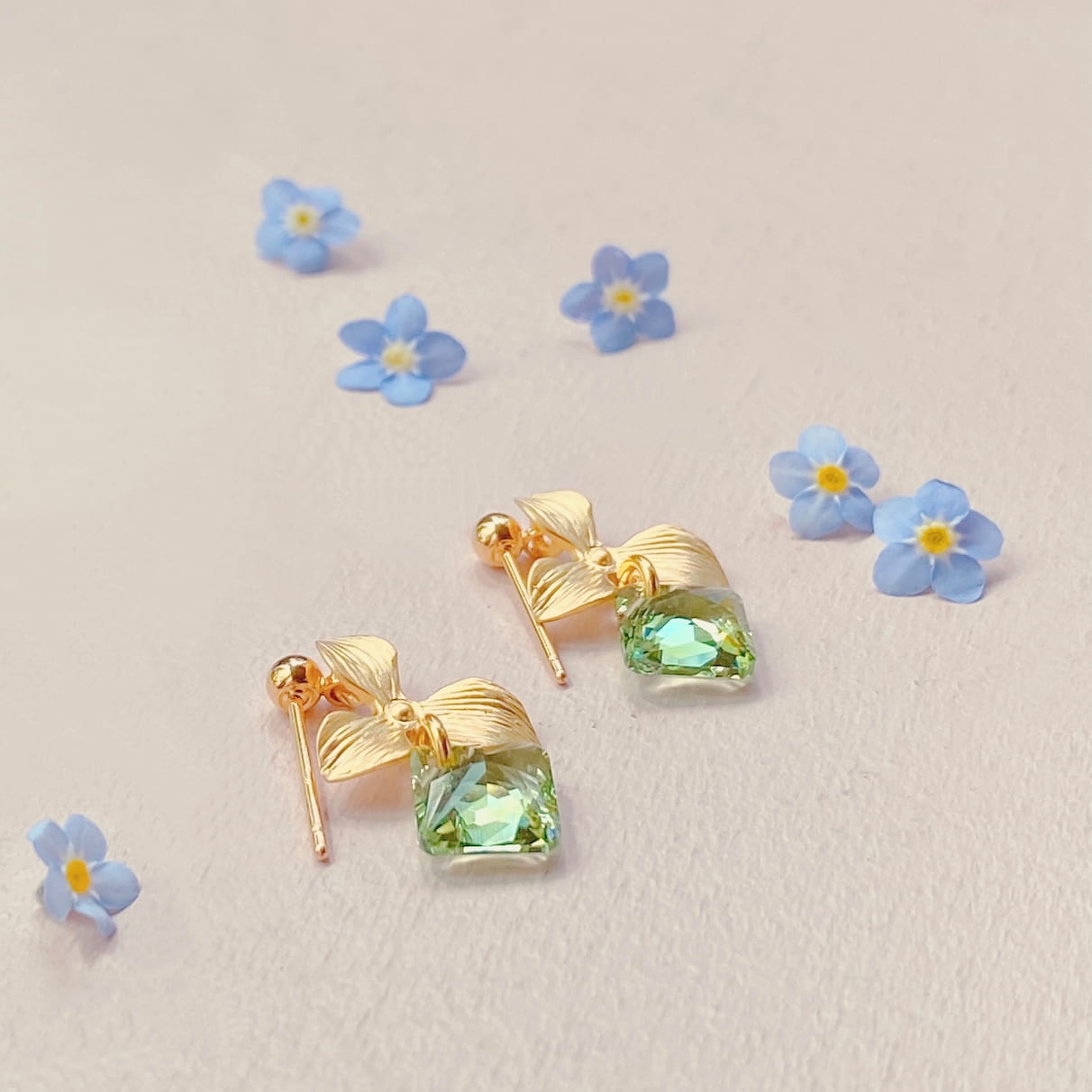 The Annette Earrings by Noémiah, featuring leaf designs and green gemstones, are showcased on a light background. Embellished with Swarovski crystals and crafted to resist tarnish, these pieces make ideal everyday earrings. A scattering of small blue flowers adds a touch of elegance to the arrangement.