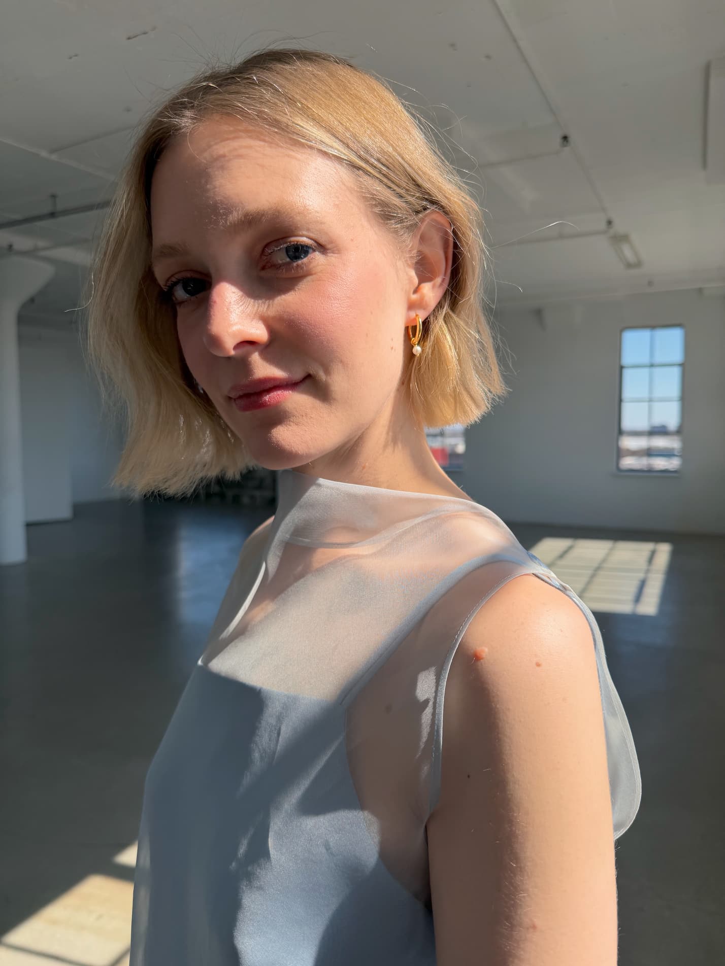 A woman with short blonde hair wears Noémiah’s Annie Earrings in a sunlit, minimalist room. She gazes gently at the camera, dressed in a sleeveless sheer top as sunlight streams through large industrial windows.