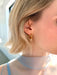 Close-up of a person with short blonde hair wearing the Annie Earrings by Noémiah—gold-plated hoops featuring a small freshwater pearl drop. A softly lit background and sheer light blue top highlight this elegant handmade accessory.