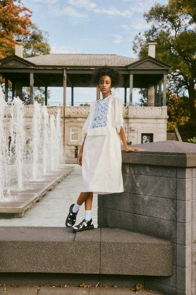 A woman in a Noémiah Ari Dress, showcasing a blue-patterned vest, leans against a stone railing near a fountain. She is wearing black sandals with white socks. The scene unfolds against the backdrop of a pavilion and autumn trees under a cloudy sky, creating an exquisite veil of breathtaking beauty.