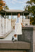 A woman in a Noémiah Ari Dress, showcasing a blue-patterned vest, leans against a stone railing near a fountain. She is wearing black sandals with white socks. The scene unfolds against the backdrop of a pavilion and autumn trees under a cloudy sky, creating an exquisite veil of breathtaking beauty.