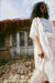 A woman in a Noémiah's Ari Dress, showcasing blue patterns on delicate voile, stands on a cobblestone path. She carries a bag adorned with a silk floral jacquard design and wears sunglasses. In the background, an old brick building with a rustic door and ivy is set against a partly cloudy sky.