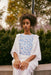 A person with curly hair is seated outdoors, wearing a Noémiah Ari Dress in white with blue floral designs. They are surrounded by greenery and lavender plants, with trees in the background, creating an autumnal atmosphere.