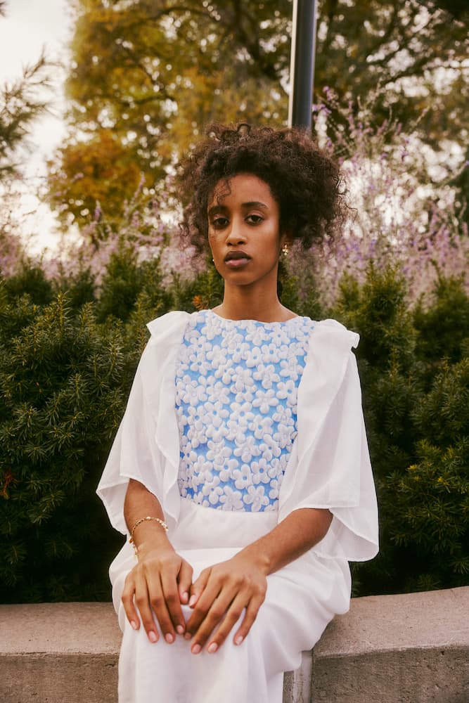 A person with curly hair is seated outdoors, wearing a Noémiah Ari Dress in white with blue floral designs. They are surrounded by greenery and lavender plants, with trees in the background, creating an autumnal atmosphere.