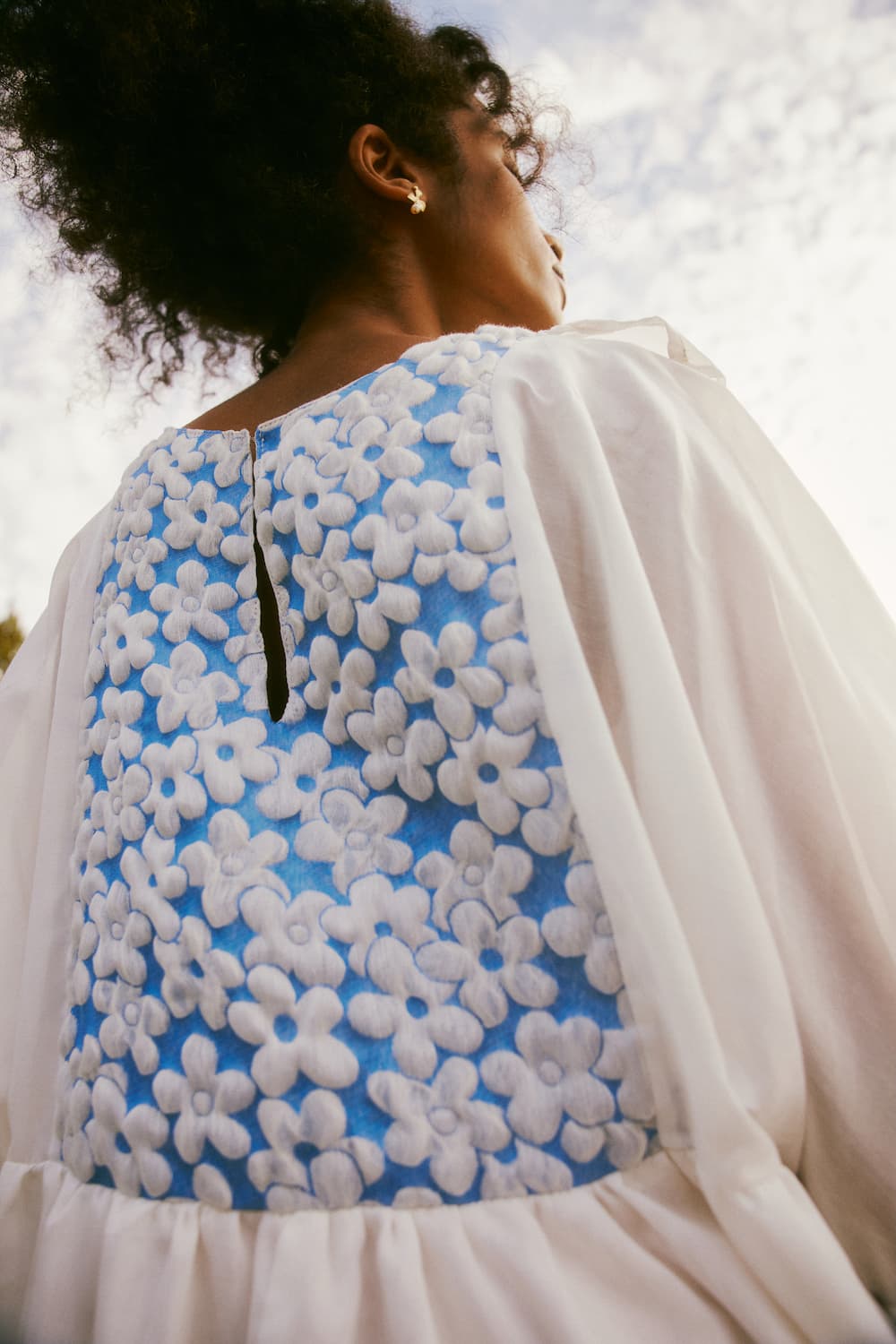 Dressed in the Ari Dress by Noémiah, a person is outdoors, seen from below in a white ensemble showcasing a back panel adorned with a blue and white floral pattern, reminiscent of delicate voile. The sky above is partly cloudy.