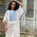 A person with curly hair wearing sunglasses and a Noémiah Ari dress, crafted in delicate voile featuring a blue floral design, stands in front of a rustic wooden door. They have one hand on their head and are looking to the side.