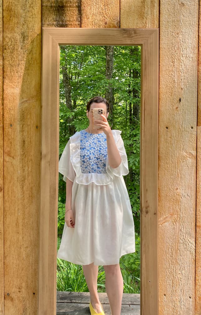 A person wearing the delicate Ari Dress by Noémiah, featuring blue floral patterns in a light voile fabric, takes a mirror selfie outdoors. The wooden-framed mirror reflects lush green trees in the background, enhancing the serene setting.