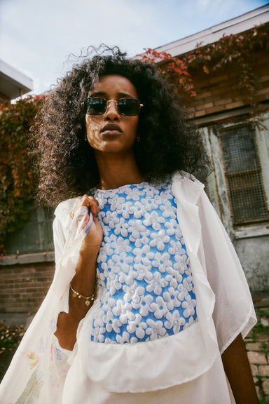 A person with curly hair and sunglasses stands confidently outdoors, wearing a Noémiah Ari Dress featuring a white and blue floral pattern. The background showcases a brick building draped in green and red ivy.