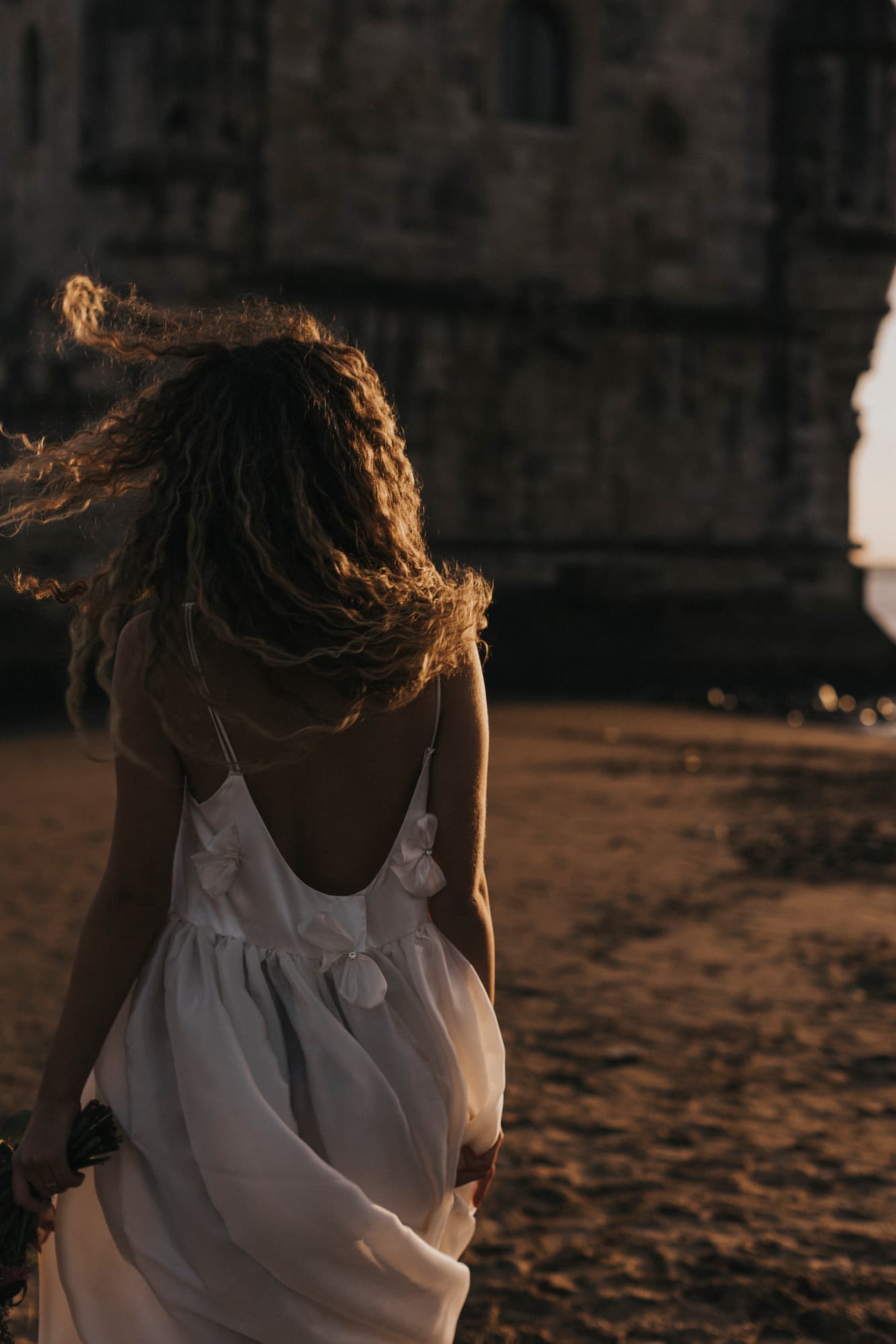 A woman with curly hair, wearing the Ariel Dress by Noémiah—a flowing silk organza creation—walks towards a stone building on a sandy beach at sunset. The scene is backlit, casting her and the structure in silhouette as gentle waves lap the shore.