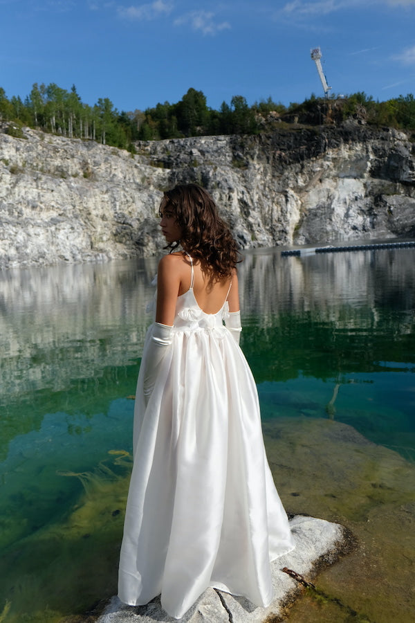 Wearing the Ariel Dress by Noémiah, a woman in a white sleeveless gown embellished with Swarovski crystals stands on a rocky edge near a clear green lake. The landscape is surrounded by cliffs and trees under a blue sky, and she has long curly hair and wears long white gloves as she gazes at the scenic view.