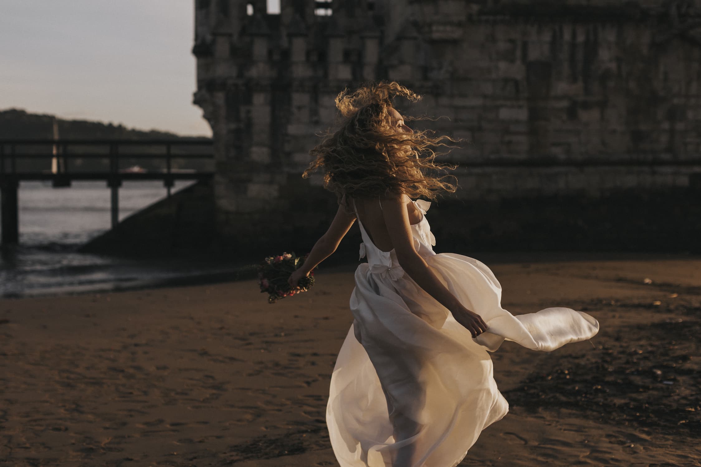 A woman in the Ariel Dress by Noémiah, crafted from flowing silk organza and embellished with Swarovski crystals, joyfully runs along a sandy beach at sunset. She clutches a small bouquet of flowers, her hair tousled by the wind, with an ancient stone structure standing majestically in the background.