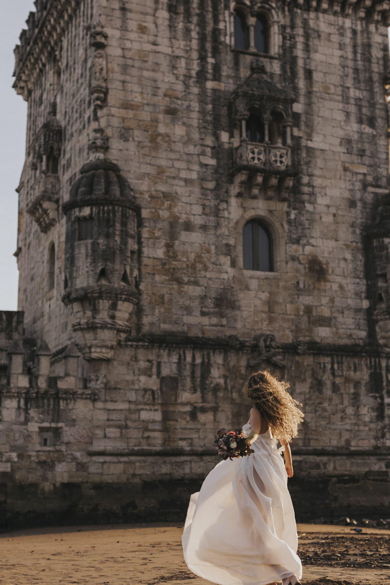 A woman with long curly hair in the Ariel Dress by Noémiah, shimmering with Swarovski crystal embellishments, holds a bouquet of flowers while standing in front of a historic stone castle. The sunlit scene highlights the castle's intricate architecture.