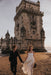 A couple strolls hand in hand on a sandy beach near the historic Belém Tower. The man wears a black suit, while the woman dazzles in an Ariel Dress by Noémiah, crafted from flowing silk organza and adorned with Swarovski crystal embellishments. She holds a bouquet as the sun casts a warm glow over the picturesque scene.