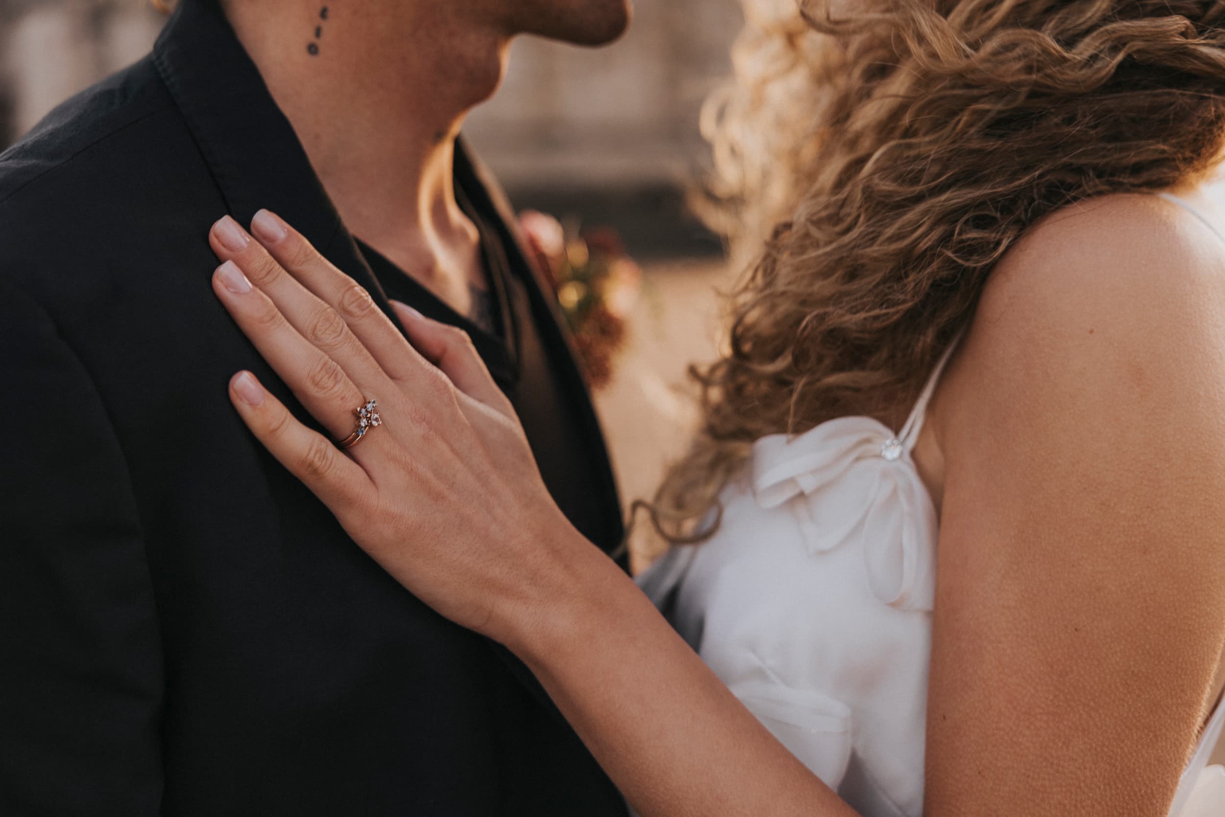 A woman with curly hair in the Ariel Dress by Noémiah, featuring silk organza and Swarovski crystal embellishments, gently places her hand, showcasing a large ring, on a man's chest. The man is wearing a dark jacket, and the image captures a romantic moment between them.