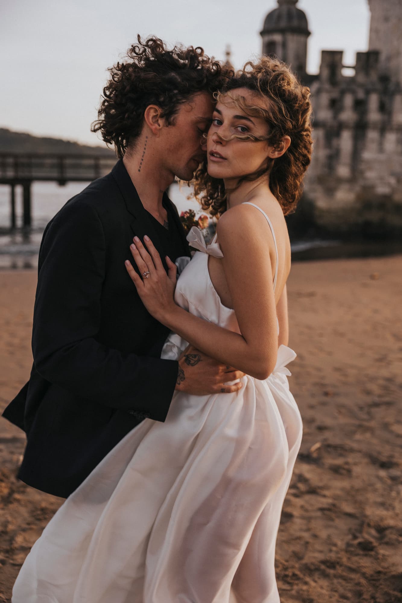 A couple embraces on a sandy beach near the ocean. The woman, wearing the Ariel Dress by Noémiah, looks towards the camera while the man in a dark suit stands close to her. A stone building and pier are visible in the background.