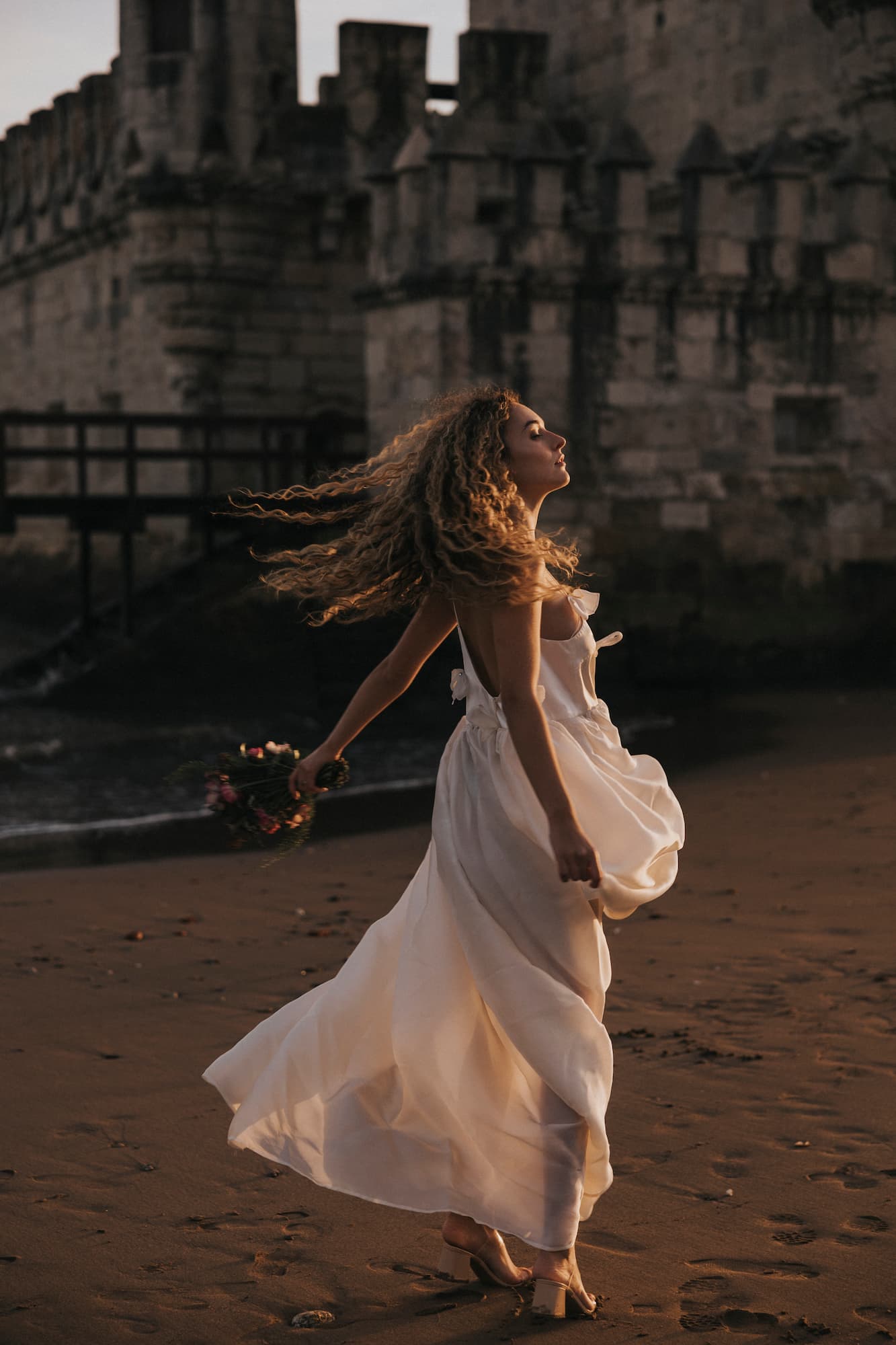 A woman in the Ariel Dress by Noémiah walks barefoot on a sandy beach at sunset, near a stone castle. She carries a small bouquet, and her curly hair streams behind her in the breeze. The customizable size ensures perfect elegance and grace with every step.