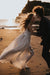 A couple joyfully dances on a beach at sunset. The woman, in the Ariel Dress by Noémiah, crafted from flowing silk organza and adorned with Swarovski crystal embellishments, has windblown hair. Meanwhile, the man is wearing a dark suit. The soft, golden light casts long shadows on the sand, with the ocean in the background.