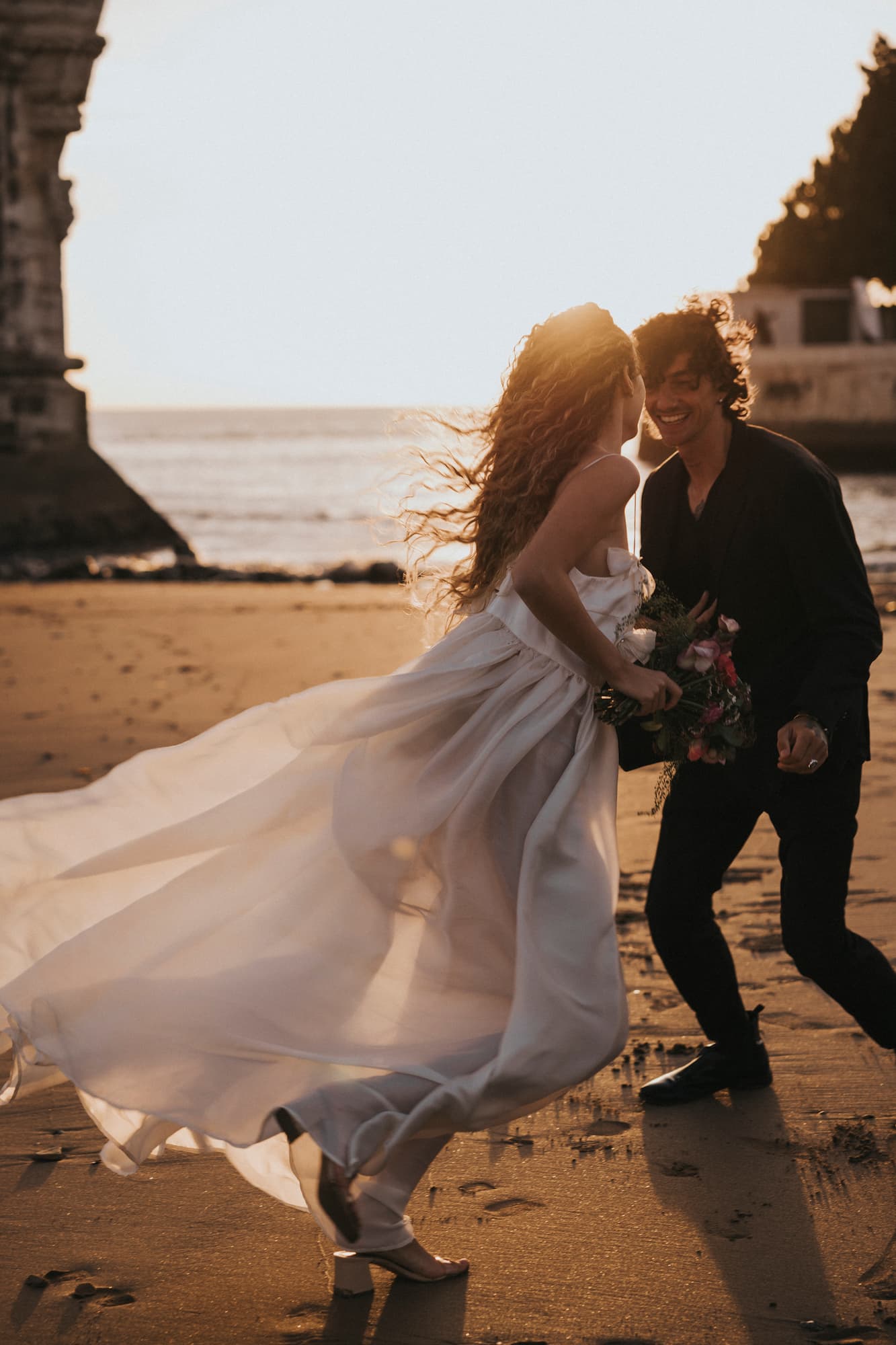 A couple joyfully runs on a beach at sunset as the woman’s Ariel Dress from Noémiah flows elegantly behind her, adorned with Swarovski crystal embellishments, while she holds a bouquet of flowers. The sea and a stone archway are visible in the background, bathed in golden light.