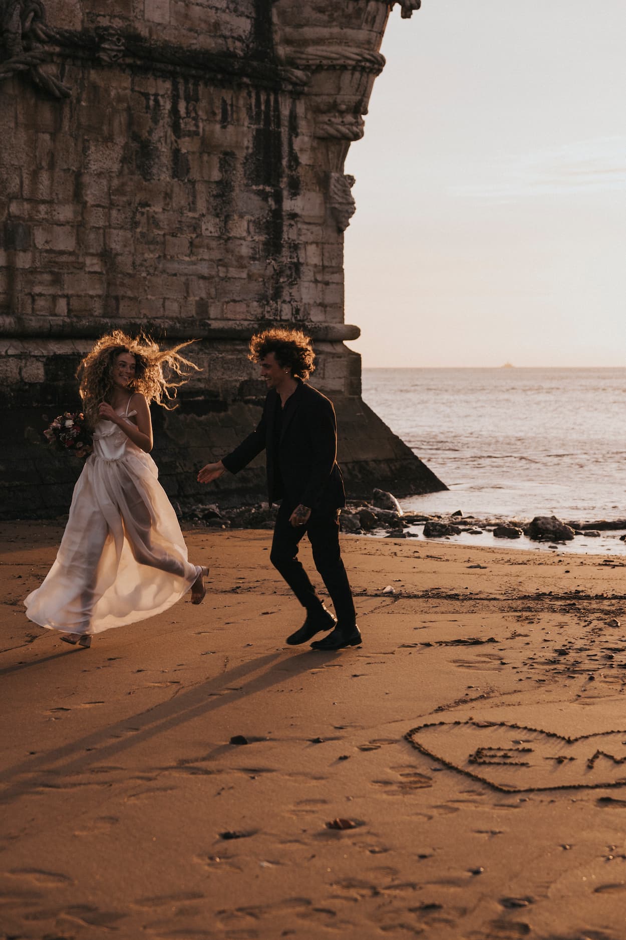 A joyful couple strolls on a sandy beach near a rocky structure. The woman, wearing the Ariel Dress by Noémiah, a flowing silk organza dress embellished with Swarovski crystals, twirls towards the man in a dark suit. The sun casts a warm glow, and a heart is drawn in the sand.