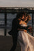 A couple embraces on a sandy beach at sunset. The man, dressed in a black suit, kisses the smiling woman on the cheek. She wears the exquisite Ariel Dress by Noémiah, featuring a silk organza fabric adorned with Swarovski crystal embellishments, while holding a bouquet of flowers. The ocean and a wooden pier are visible in the background.