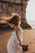 A person with curly hair spins joyfully on a beach, wearing the Ariel Dress by Noémiah, a flowing silk organza adorned with Swarovski crystal embellishments. They hold a bouquet of flowers, and a rocky cliff is visible in the background as the sun sets over the calm ocean, casting a warm glow.