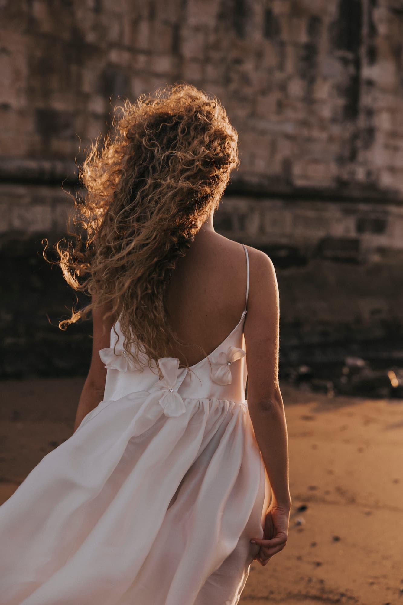 A person with long curly hair, wearing the Ariel Dress by Noémiah—a stunning white silk organza dress embellished with Swarovski crystals and a delicate bow—stands on a beach at sunset. A stone wall forms the backdrop as the dress flows gracefully in the wind, bathed in the warm glow of the sun.