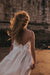 A person with long curly hair, wearing the Ariel Dress by Noémiah—a stunning white silk organza dress embellished with Swarovski crystals and a delicate bow—stands on a beach at sunset. A stone wall forms the backdrop as the dress flows gracefully in the wind, bathed in the warm glow of the sun.