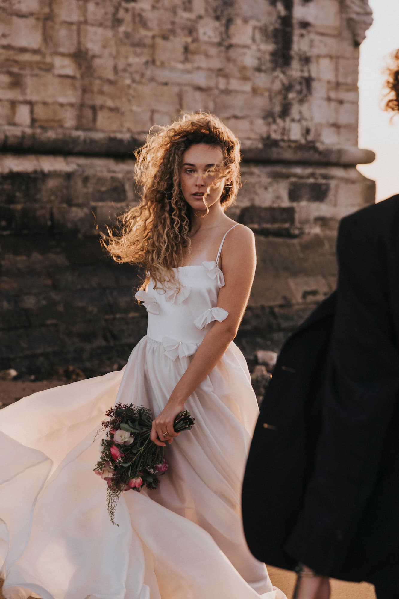 A woman with curly hair wears the Ariel Dress by Noémiah, its white flowing design gracefully capturing the wind. She holds a bouquet of flowers and stands near a stone wall, while a blurred figure in dark clothing partially appears in the foreground, adding an element of mystery to the serene scene.