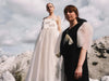 Under a cloudy sky, two women stand on rocky terrain. One wears the flowing Ariel Dress by Noémiah in white silk organza, while the other is dressed in a black version featuring puffy white sleeves. Both dresses are decorated with elegant bow details and subtle Swarovski crystal embellishments, creating a serene and ethereal atmosphere.