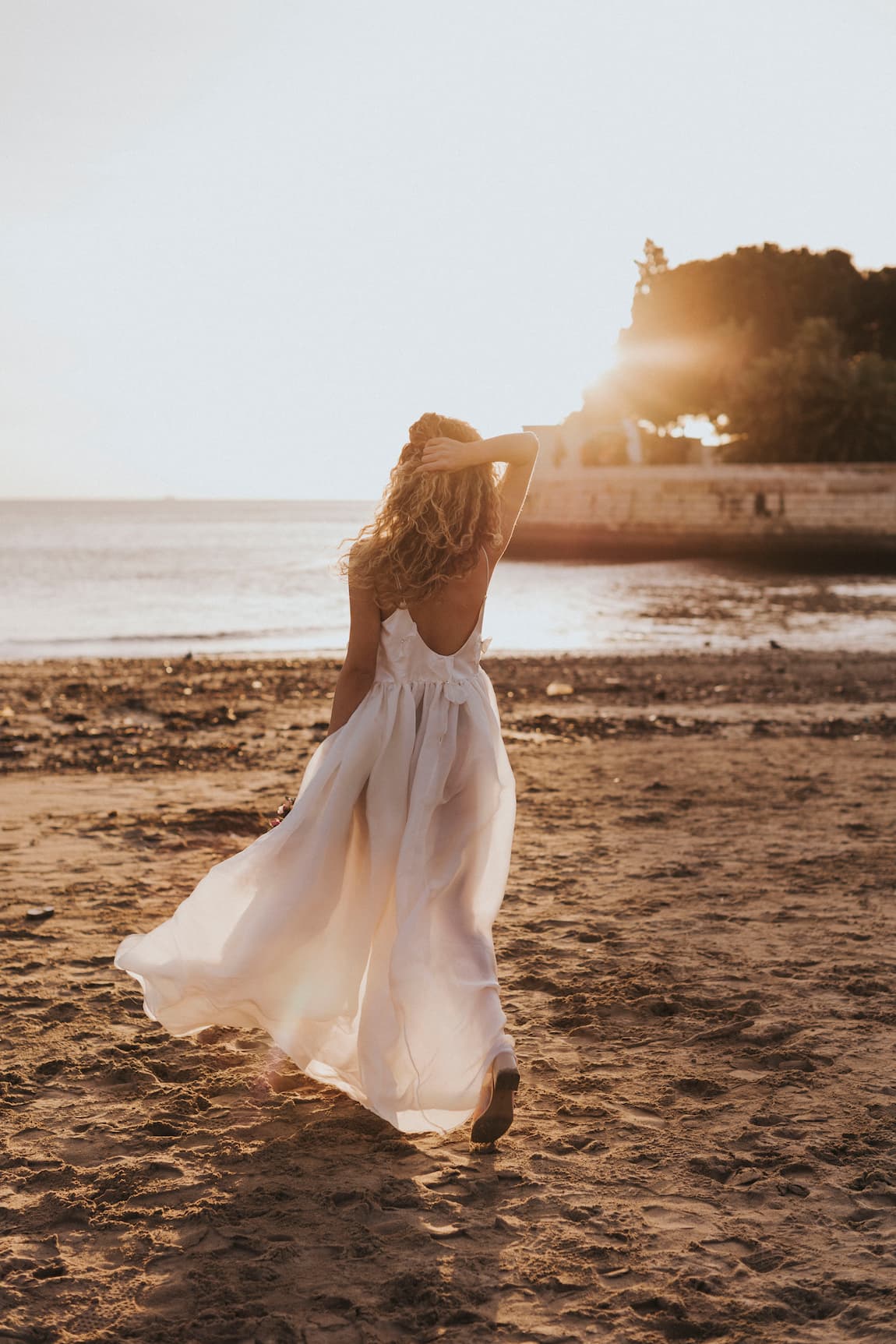 A woman wearing the Ariel Dress by Noémiah strolls along a sandy beach at sunset. Her long, wavy hair cascades around her as she gently touches her head while facing the ocean. The sun's warm light dances off the Swarovski crystal embellishments on her dress, enveloping the scene in a dreamy glow.