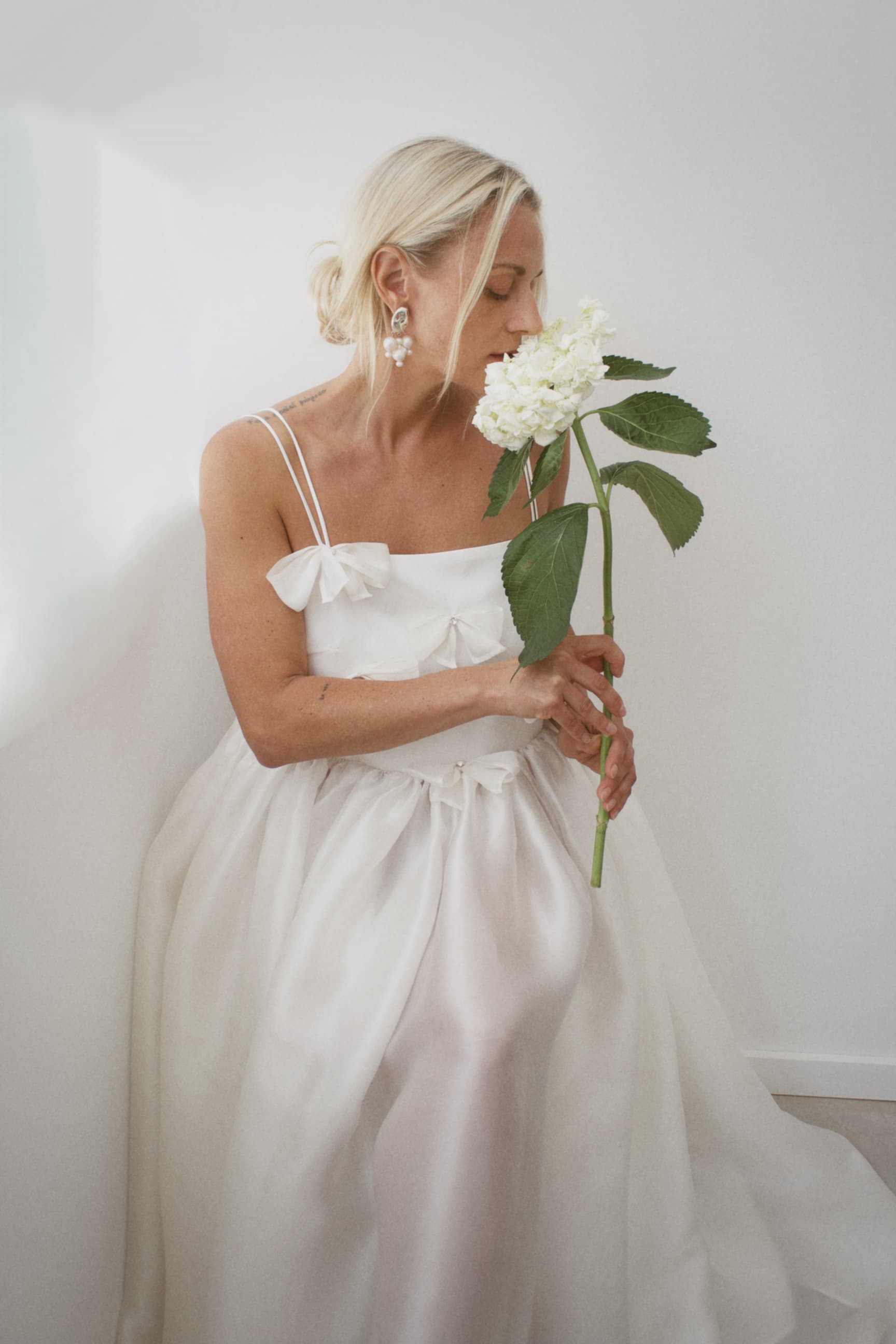 Wearing the Ariel Dress by Noémiah, a woman sits gracefully against a white wall, holding a white flower near her face. Her hair is styled in a low bun, and she accessorizes with dangling earrings that feature Swarovski crystal embellishments. The scene exudes softness and serenity, embodying timeless elegance.