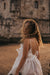 A woman with curly hair wearing the Ariel Dress by Noémiah, a silk organza creation adorned with Swarovski crystals, stands on a sandy beach, looking back over her shoulder. The wind gently blows her hair across her face, while a stone building in the background is softly lit by the sun.