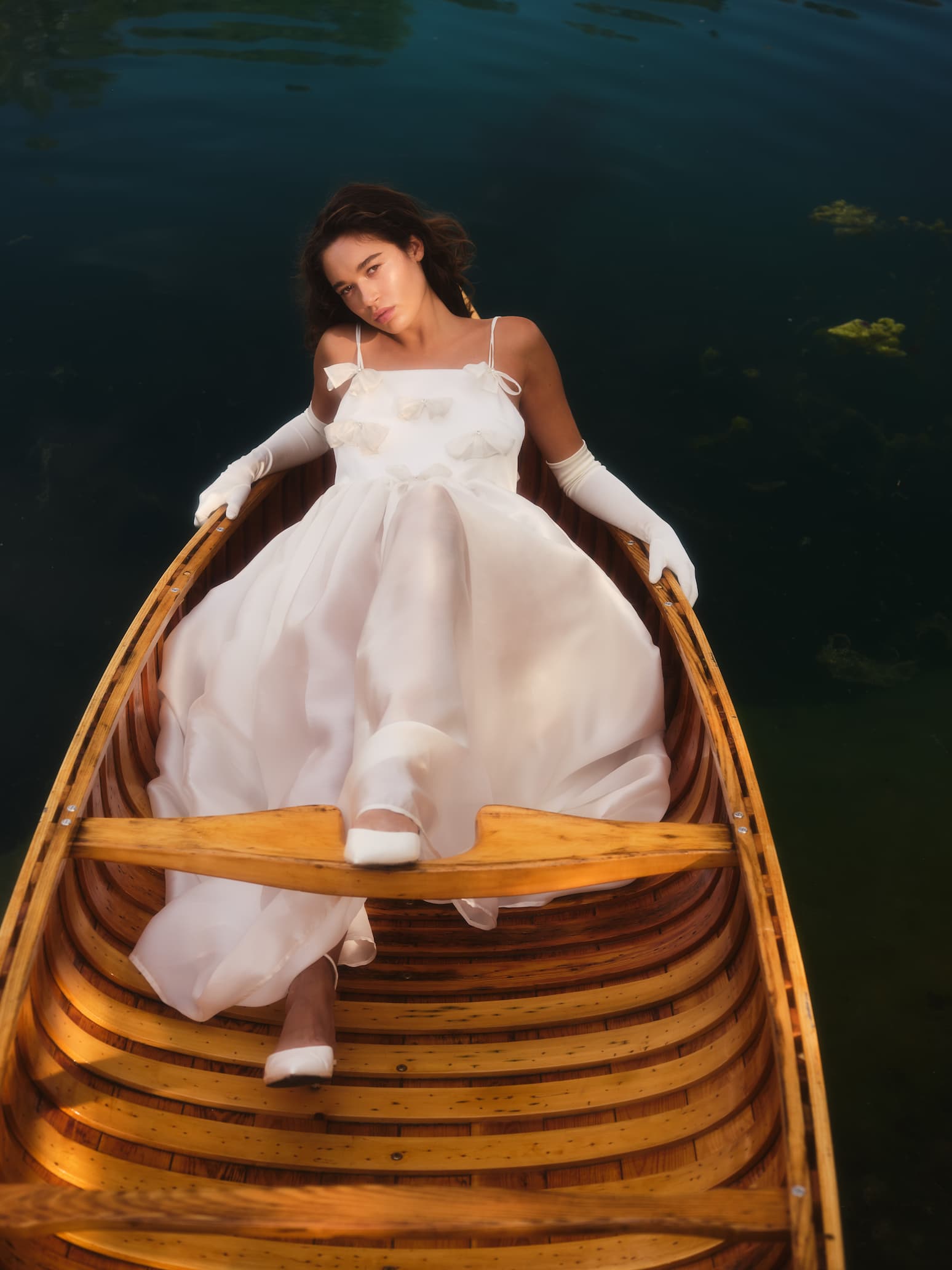 A woman in the Ariel Dress by Noémiah, crafted from flowing silk organza and adorned with Swarovski crystal embellishments, lies elegantly in a wooden boat on a calm lake. The water is dark and reflective, contributing to the scene's serene and peaceful atmosphere.