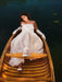 A woman in the Ariel Dress by Noémiah, crafted from flowing silk organza and adorned with Swarovski crystal embellishments, lies elegantly in a wooden boat on a calm lake. The water is dark and reflective, contributing to the scene's serene and peaceful atmosphere.