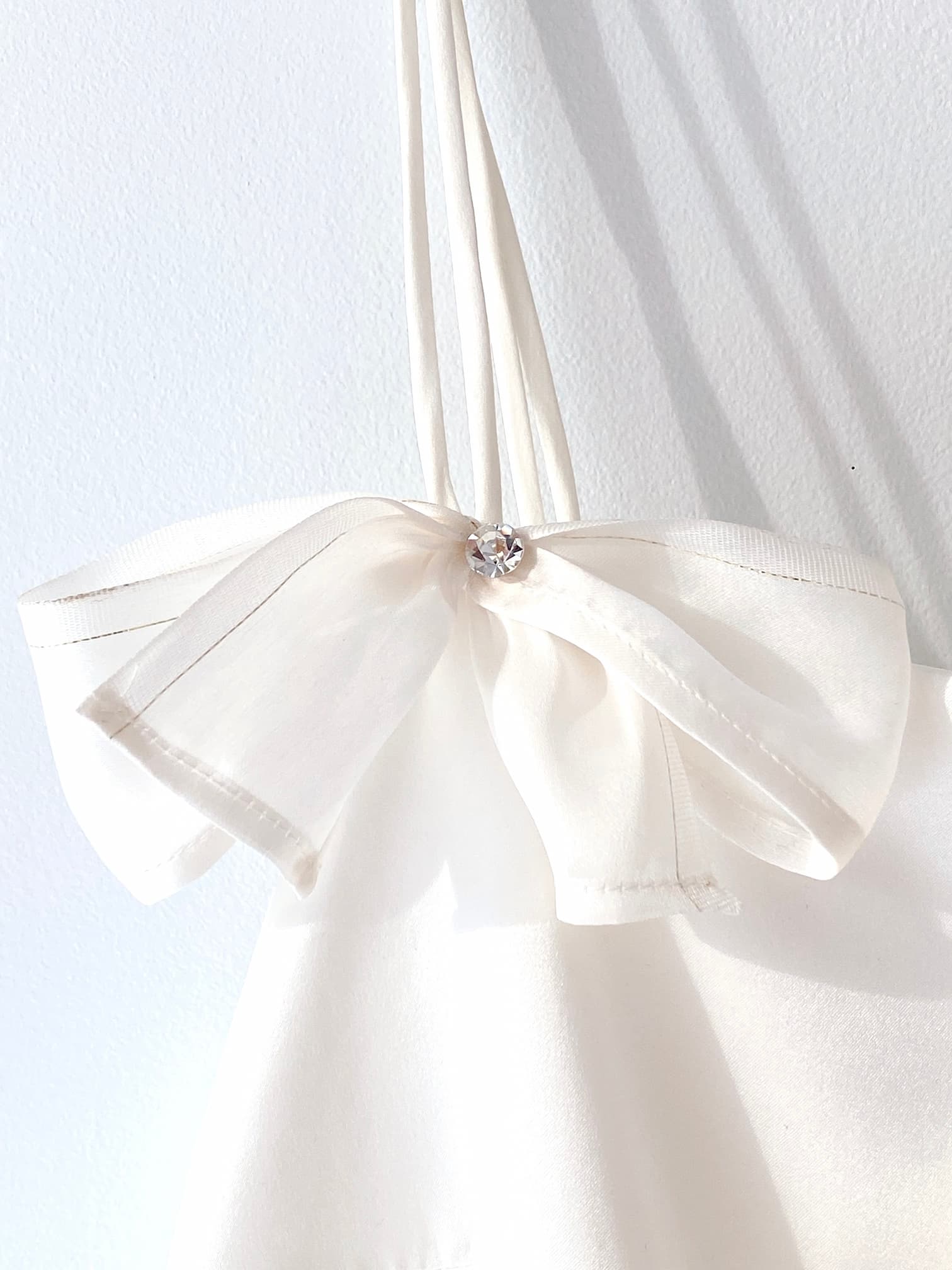 A white fabric bag, featuring a delicate, sheer white bow adorned with Swarovski crystal embellishments at its center, hangs against a white wall. This elegant bow adds a touch of sparkle and sophistication to the minimalist design of the Ariel Dress by Noémiah.