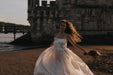 A woman in the Ariel Dress by Noémiah, crafted from silk organza and adorned with Swarovski crystal embellishments, holds a bouquet of flowers on a sandy beach at sunset. Her curly hair is windswept, and a historic stone building in the background casts gentle shadows.